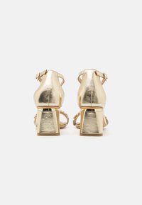 Las sandalias de tacón metálico dorado cuentan con un tacón cuadrado, una correa para el tobillo con hebilla y un diseño de punta abierta. Textura suave con acentos decorativos.