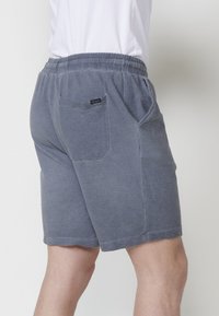 Uomo che indossa pantaloni corti grigi a trama con vita elastica e un piccolo etichetta nera sulla tasca posteriore, abbinati a una camicia bianca.