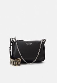 Fekete bőr keresztpántos táska texturált felülettel, ezüst színű láncpánttal és egy kontrasztos bézs színű anyagpánttal, amely fekete betűkkel van díszítve.