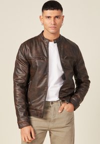 Giacca in finta pelle marrone con colletto rotondo, zip frontale, due tasche sul petto con zip, indossata su una maglietta bianca e pantaloni beige.