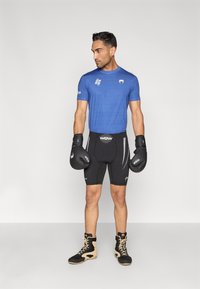 T-shirt azul desportivo com padrão geométrico texturizado, combinado com calções de compressão pretos. Luvas de boxe pretas e calçado preto e castanho completam o visual.