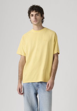 Joven de pie vistiendo una camiseta amarilla lisa de manga corta y vaqueros azul claro frente a un fondo claro y liso.