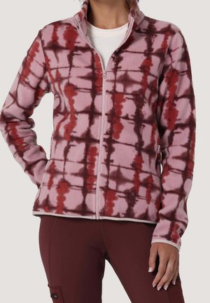 Frau trägt eine pink-rote Batik-Fleecejacke mit Frontreißverschluss, kombiniert mit dunkelroten Hosen und einem weißen Unterhemd.