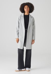 Lichtgrijze wollen mix jas met een reverskraag, een enkele knoopsluiting en lange mouwen; gecombineerd met donkere jeans en witte sneakers.