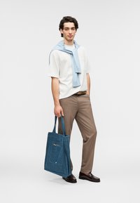 Τσάντα tote καμβά σε μπλε χρώμα με τσέπη με φερμουάρ, που φοράει ένα μοντέλο με λευκό πουκάμισο και καφέ παντελόνι, κρατώντας την τσάντα από τα χερούλια της.