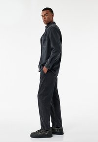 Schwarze Jeanshemd mit einer Brusttasche, kombiniert mit lockeren schwarzen Hosen und strukturierten schwarzen Sneakers, vor einem neutralen Hintergrund präsentiert.