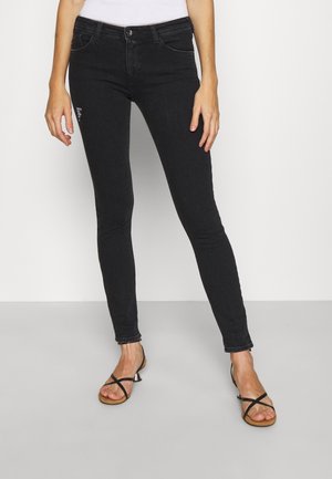 Jean noir slim porté par une personne debout, associé à des talons noirs ouverts à brides et un haut blanc partiellement visible.