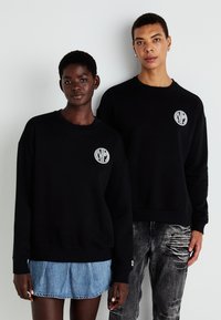 Sorte hettegensere har et rundt logo-patch på venstre bryst. Modellen til venstre har på seg et denimskjørt; den til høyre har slitte jeans.