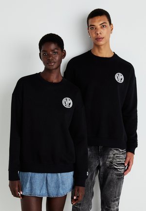 Sorte hettegensere har et rundt logo-patch på venstre bryst. Modellen til venstre har på seg et denimskjørt; den til høyre har slitte jeans.