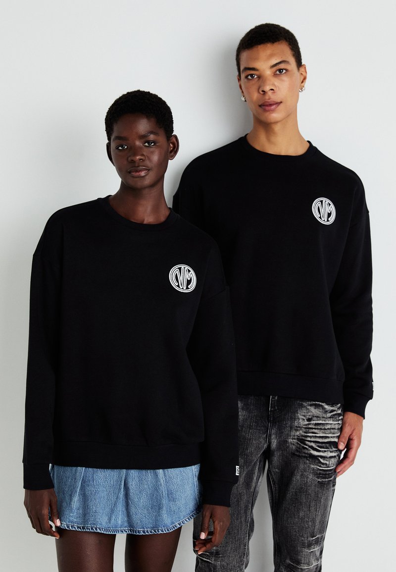 Sorte hettegensere har et rundt logo-patch på venstre bryst. Modellen til venstre har på seg et denimskjørt; den til høyre har slitte jeans.