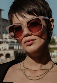 Katzenaugen-Sonnenbrille mit einem durchsichtigen, hellrosa Rahmen und dunklen roten Gläsern, getragen mit geschichteten goldenen Halsketten, vor einem unscharfen Hintergrund.