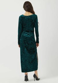 Dlouhá tyrkysová sametová šaty s dlouhými rukávy, tečkovaným vzorem, bočním rozparkem a zaobleným výstřihem; kombinováno s černými vysokými podpatky.