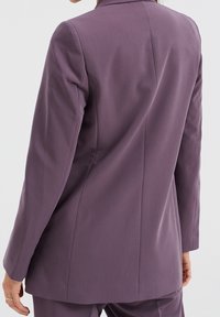 Skräddarsydd lila blazern med minimalistisk design, strukturerade axlar, en sprund bak och mjuk tygtextur.