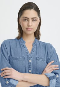 Chemise en denim avec un col en V, deux poches poitrine, fermeture boutonnée sur le devant et manches retroussées. Couleur bleu clair avec une texture douce.