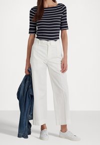 Vita vida byxor med hög midja, bälte och fickor, i kombination med en marinblå och vit randig kortärmad topp. Denimjacka hängande över armen, vita sneakers.