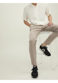 Chemise à manches courtes de couleur claire, pantalon beige slim fit, et baskets noires avec des accents blancs, assis sur un tabouret blanc contre un fond clair.