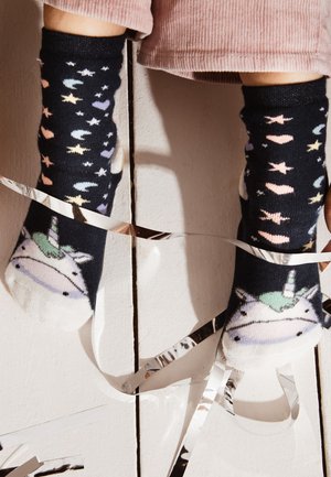 Feet wearing black socks with unicorn faces, stars, hearts, and moons, standing on white wooden floor with silver ribbon strands.