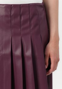 Fialová plisová kožená sukně s hladkým povrchem, obsahující několik vertikálních záhybů a boční zip. Ruka spočívá na pasu.