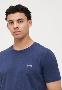 Tmavě modré tričko s krátkým rukávem vyrobené z jemné tkaniny. Má kulatý výstřih a nenápadné logo "BOSS" v bílé barvě na hrudi.
