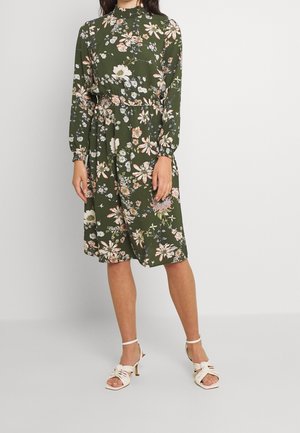 Nainen, joka pukeutunut polvipituiseen oliivinvihreään mekkoon, jossa on kukkakuvio ja pitkät hihat, yhdistettynä valkoisiin korkokenkiin.