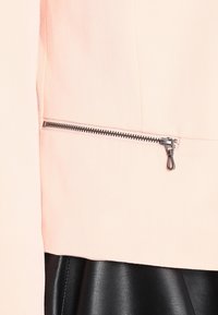 Ljusrosa jacka i tyg med en siddragkedja i silverfärgad hårdvara, med en slät textur. Svart material är synligt under jackan.