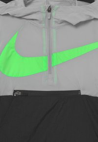Celkový pohled na šedou a černou bundu s zeleným logem Nike swoosh a viditelnými zipy na předních kapsách a límci.