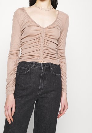 Femme portant un haut beige froncé à manches longues avec un décolleté en V profond, associé à un jean noir taille haute sur fond blanc.