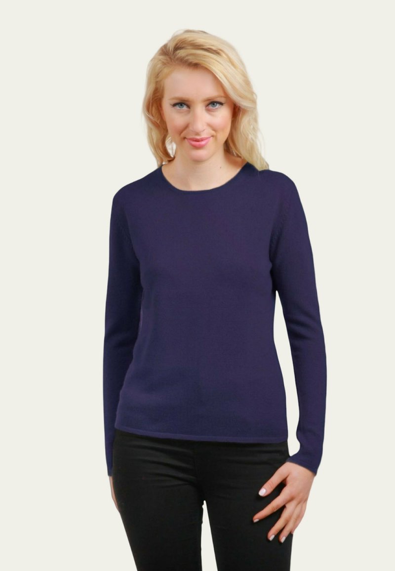 Femme blonde portant un pull simple à manches longues bleu foncé et un pantalon noir, se tenant devant un fond clair avec un léger sourire.