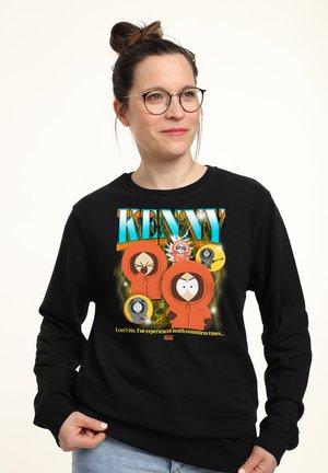 Kvinde med briller iført en sort sweatshirt med flere tegneseriefigurer i orange hætter og tekst om Kenny fra South Park.