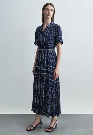 Kvinde stående iført en navyblå midi-kjole med hvide paisley- og prikmønstre, korte ærmer, knapper foran og sorte strop-sandaler.