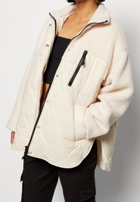 Krämfärgad jacka med kviltad design, utrustad med en fluffig krage, en ficka med dragkedja på bröstet och överdimensionerade ärmar.