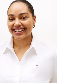 Λευκό πουκάμισο με γιακά, κατασκευασμένο από λεία υφή, με μικρό, κεντημένο λογότυπο στο αριστερό στήθος. Η επαφή με το δέρμα δείχνει φυσική υφή.