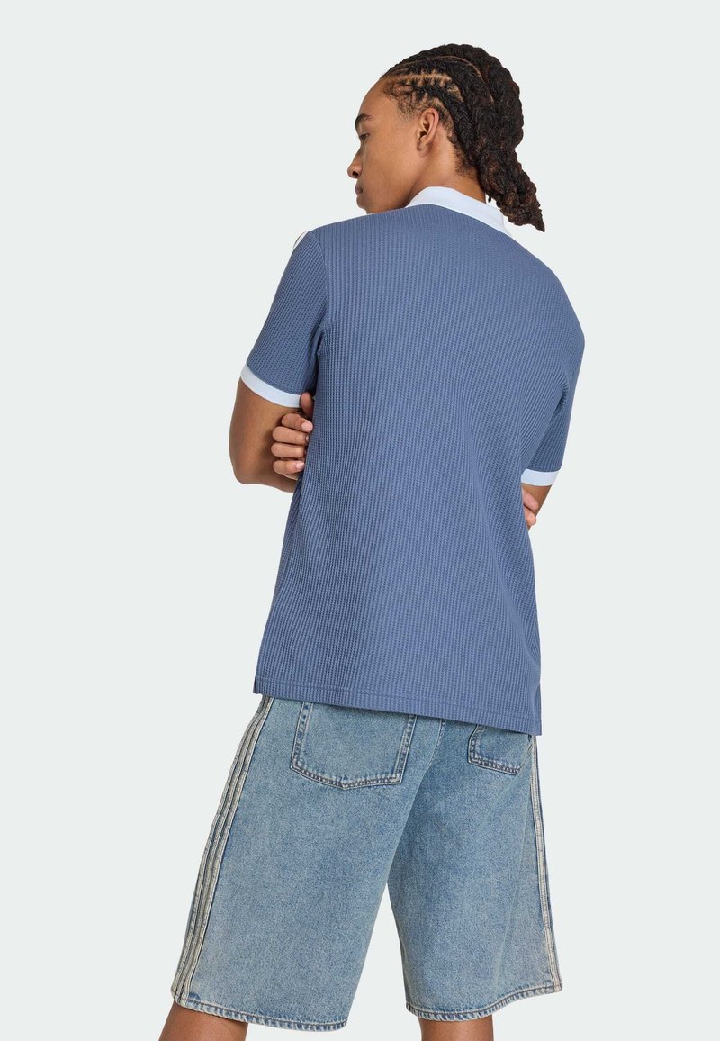 Persona con capelli intrecciati che indossa una camicia blu a maniche corte con collo e polsini bianchi, e pantaloncini di jeans larghi con strisce laterali.