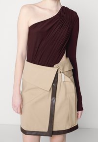 Femme portant un top marron à une épaule avec manches longues et une jupe portefeuille beige avec bordure foncée et boucle de ceinture à la taille.