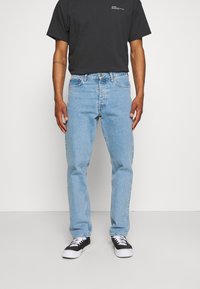 Vaqueros de mezclilla azul claro con corte recto, cinco bolsillos y remaches de metal. Combinados con una camiseta negra de manga corta y zapatillas negras.