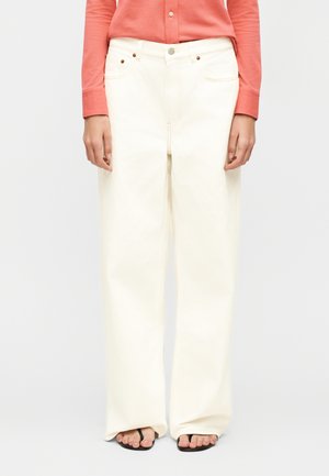 Woman wearing high-waist wide-leg white pants, coral long-sleeve buttoned shirt, and black open-toe sandals standing against a plain background.