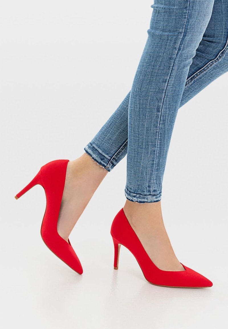 Rode suède hoge hakken met een spitse neus en een 10 cm stiletto hak; gedragen met lichtblauwe, strakke denim jeans met rafelige zomen.