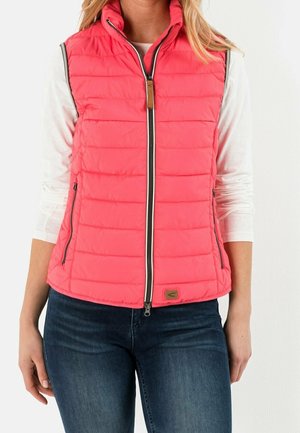 Vrouw die een fel koraal gekleurd doorgestikt vest draagt met een ritssluiting aan de voorkant en ritszakken over een wit shirt met lange mouwen en blauwe spijkerbroek.