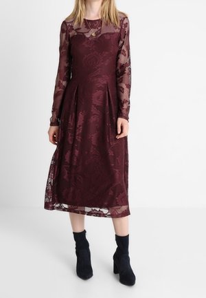 Femme portant une robe midi en dentelle bordeaux à manches longues avec un motif floral et des bottines noires, debout devant un fond blanc.