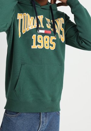 Persoon draagt een groene hoodie met de tekst "Tommy Jeans 1985" in geel en wit, gecombineerd met een blauwe spijkerbroek.