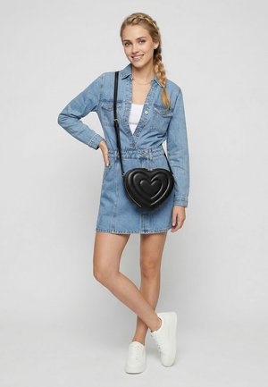 Junge Frau mit geflochtenem Haar trägt Jeanskleid, weiße Turnschuhe und eine schwarze herzförmige Umhängetasche und lächelt dabei.