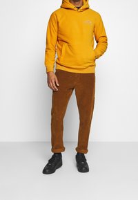 Sudadera amarilla con bolsillo frontal, combinada con pantalones de pana marrones. Zapatillas negras completan el atuendo casual.