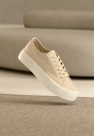 Tenis bej din pânză cu talpă groasă de cauciuc alb, așezat pe un covor bej texturat și o canapea rotundă bej în fundal.