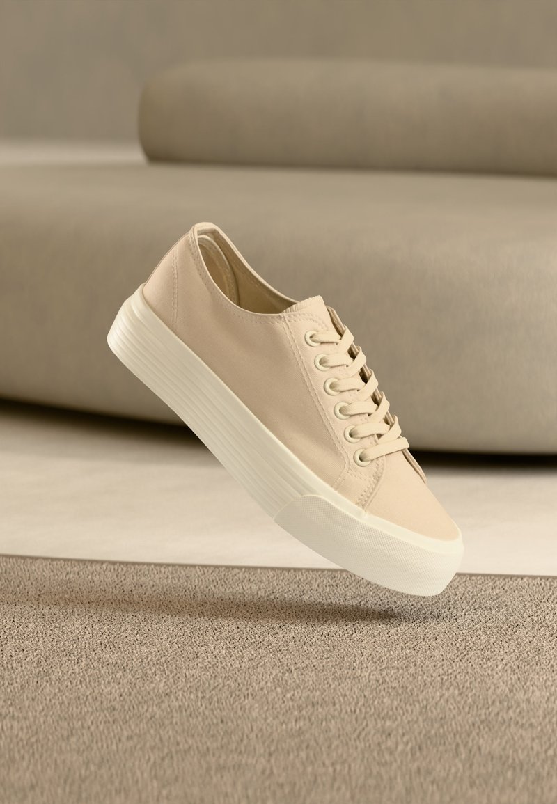 Baskets en toile beige avec semelle épaisse en caoutchouc blanc, posées sur un tapis beige texturé avec un canapé beige arrondi en arrière-plan.