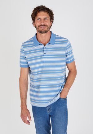 Uomo con i capelli ricci che indossa una maglietta polo a righe blu e bianche e jeans blu, in piedi con una mano in tasca.