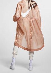 Persikofärgad, lätt och genomskinlig ytterklädsel med elastisk midja, med en framträdande vit Nike-logotyp på baksidan. Pullover-design.