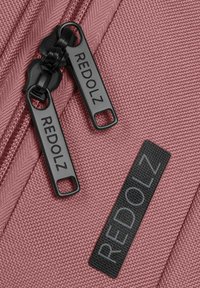 Rosa Stofftasche mit strukturierter Oberfläche, ausgestattet mit schwarzen Reißverschlüssen und Etiketten, die "REDOLZ" in Grau zeigen. Die Reißverschlüsse haben eine rechteckige Form.