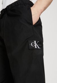 Černé joggingové kalhoty z hladké látky s elastickým pasem, postranní kapsou a výrazným textilním štítkem s logem "Calvin Klein Jeans".