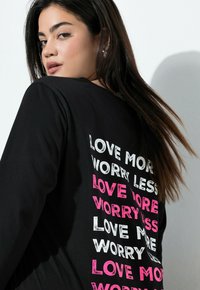Svart långärmad skjorta med djärv, färgglad text på baksidan: "ÄLSKA MER," "OROA DIG MINDRE" i vitt och rosa, med en sliten textur.