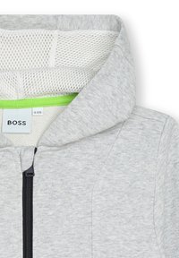 Šedá mikina na zip s prodyšnou podšívkou a živým zeleným akcentem uvnitř límce. Obsahuje černý zip a značkovou nálepku.